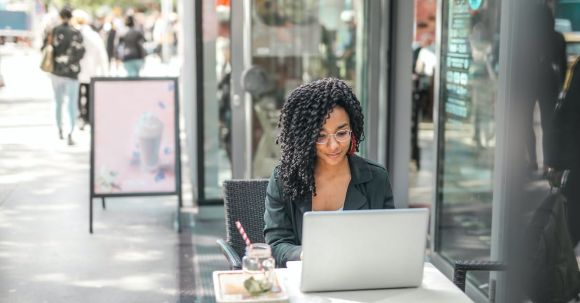 Online Business - High angle of pensive African American female freelancer in glasses and casual clothes focusing on screen and interacting with netbook while sitting at table with glass of yummy drink on cafe terrace in sunny day