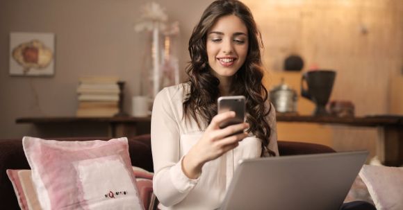 Online Business - Woman Sitting on Sofa While Looking at Phone With Laptop on Lap