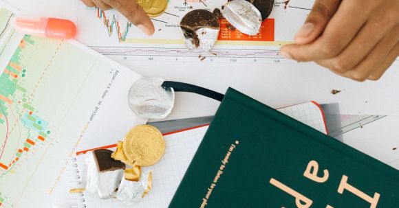 Business Management - Person Holding a Gold Chocolate Coin on the Table with a Green Book